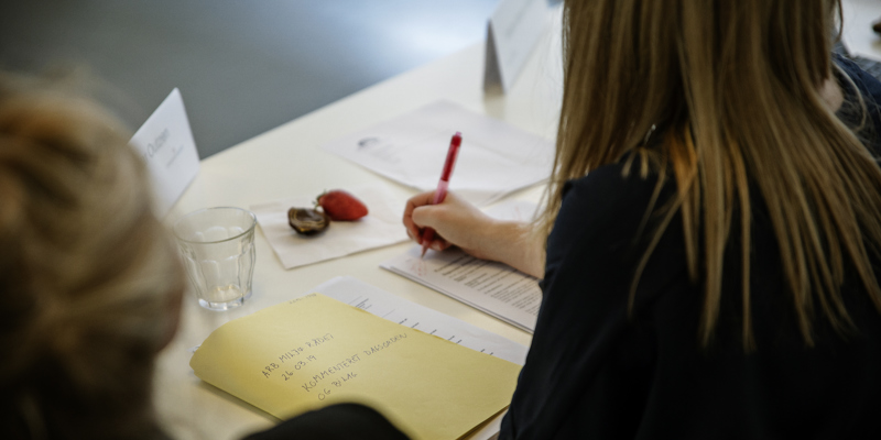 Møde i Arbejdsmiljørådet - ser en kvinde bagfra med langt brunt hår i sort bluse, som skriver på et papir