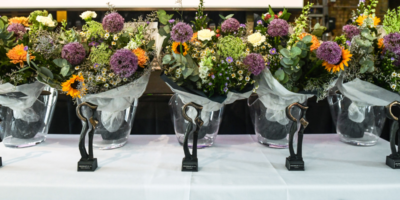 4 blomsterbuketter og 4 af Arbejdsmiljøprisens statuetter præsenteret på et bord med hvid dug.